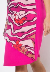 Falda rosa brillante con una superposición transparente que presenta líneas onduladas y patrones florales multicolores, llevada por una persona con una mano y piernas visibles.