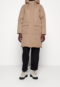 Beige, knælang parka med hætte, frontlynlås, to store sidelommer og en glat tekstur; båret over sorte bukser og beighikersko.