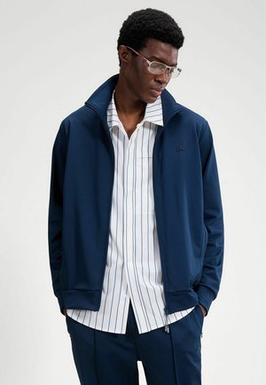 Hombre con gafas, vestido con chaqueta azul marino con cierre de cremallera, camisa blanca con rayas verticales azul marino y pantalones a juego, con las manos en los bolsillos.