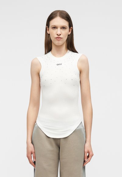 Νεαρή γυναίκα με ίσια καστανά μαλλιά, φοράει λευκό αμάνικο τοπ με μικρές μεταλλικές λεπτομέρειες και μπεζ παντελόνι, στέκεται μπροστά σε ένα απλό φόντο.