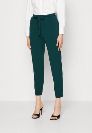 Femme portant un pantalon fuselé vert foncé avec un lien à la taille, associé à un chemisier blanc et des sandales noires à talons hauts et bouts ouverts.