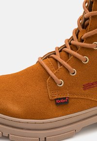Tan suede boot with round laces, metal eyelets, and a rubber sole. Features a red "Kickers" logo on the side and textured surface.