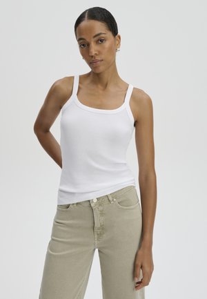 Donna con capelli scuri in un top bianco senza maniche e pantaloni beige, in piedi con una mano dietro la schiena contro uno sfondo chiaro e semplice.