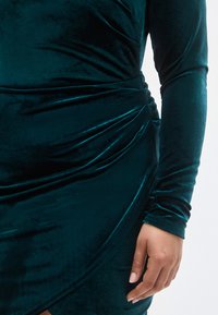 Βελούδινο φόρεμα σε τιρκουάζ απόχρωση με μακριά μανίκια, με σχεδιασμό σε πτυχές και υφή. Κοντινό πλάνο που δείχνει το απαλό ύφασμα και τις διακριτικές λεπτομέρειες στις πτυχώσεις.