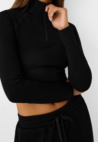 Haut noir à manches longues avec fermeture à quart de glissière, en tissu extensible, coupe ajustée et texture lisse, associé à un pantalon à taille élastiquée.