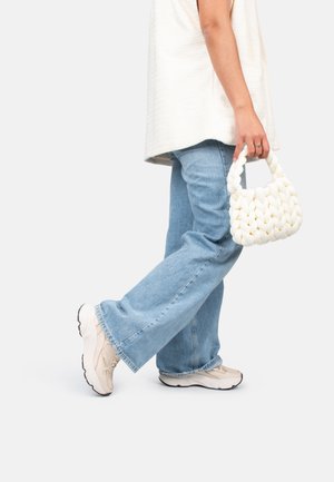 Witte geweven handtas met een robuust ontwerp, vastgehouden door een persoon in een losse beige top en lichtblauwe jeans, gecombineerd met lichte sneakers.