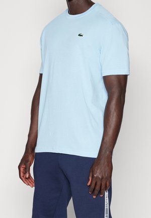 Uomo che indossa una camicia azzurra a maniche corte con piccolo logo coccodrillo verde e pantaloni blu navy con striscia bianca con logo Lacoste.