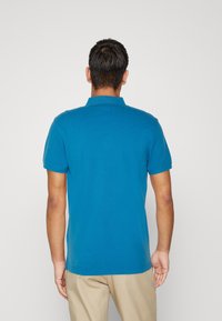 Hombre con cabello corto y oscuro, vestido con una camiseta polo azul de manga corta y pantalones beiges, visto desde atrás sobre un fondo liso.