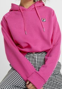 Růžová mikina s kapucí a stahovací šňůrkou, s nášivkou loga. Spárovaná s kalhotami s černobílým vzorem houndstooth. Hladká textilie.