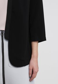 Dłoń kobiety spoczywająca na boku, ubrana w czarną kurtkę na rękawach nad różowym topem i spódnicę w kolorze jasnoszarym z wysokim stanem oraz czarnym zamkiem.