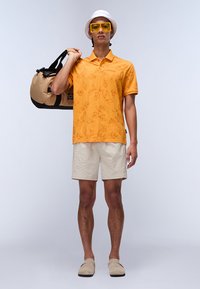 Orange polo skjorte med bladmotiver, beige shorts og lysebrune slip-on sko. Klare gule solbriller og hvid bøttehat. Sort og beige duffel bag.