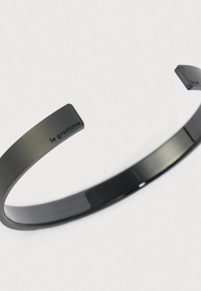Close-up de uma elegante pulseira aberta em metal escuro com "le gramme" gravado em uma das extremidades, contra um fundo claro e simples.
