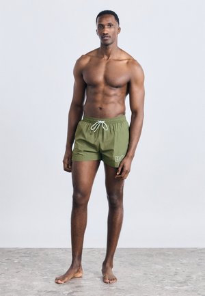 Athletischer Mann steht barfuß auf einem grauen Boden, trägt olivgrüne Badeshorts mit weißem Kordelzug und "BOSS"-Logo am linken Bein.