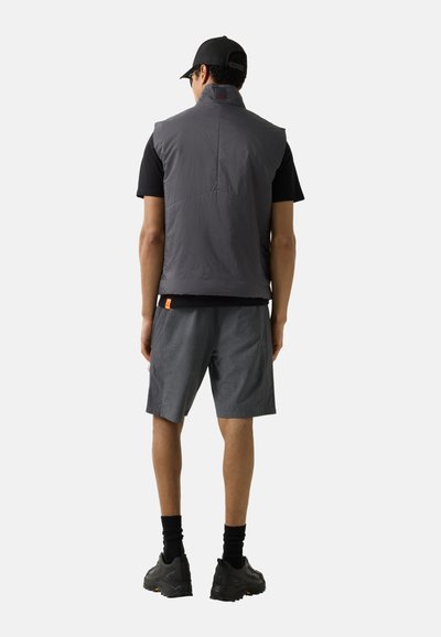 Man die met zijn rug naar de camera staat, draagt een zwarte pet, een grijs mouwloos vest, een zwart t-shirt, grijze korte broek, zwarte sokken en zwarte sportschoenen tegen een witte achtergrond.