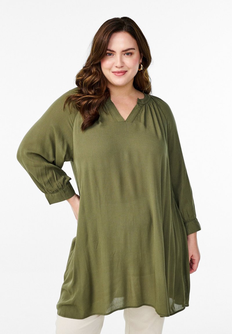 Olivengrünes Longsleeve-Tunika mit V-Ausschnitt, gerafftem Ausschnitt und lockerer Passform, kombiniert mit hellen Unterteilen. Texturierte Stoff.
