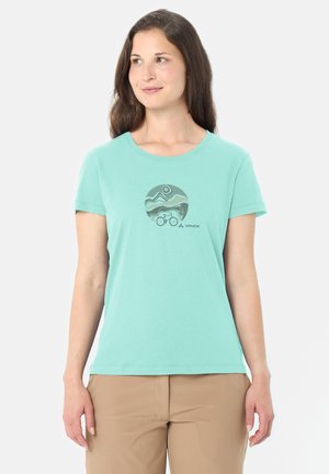 Donna che indossa una t-shirt verde menta con grafica di montagna e bicicletta, abbinata a pantaloni beige, in piedi contro uno sfondo bianco uniforme.