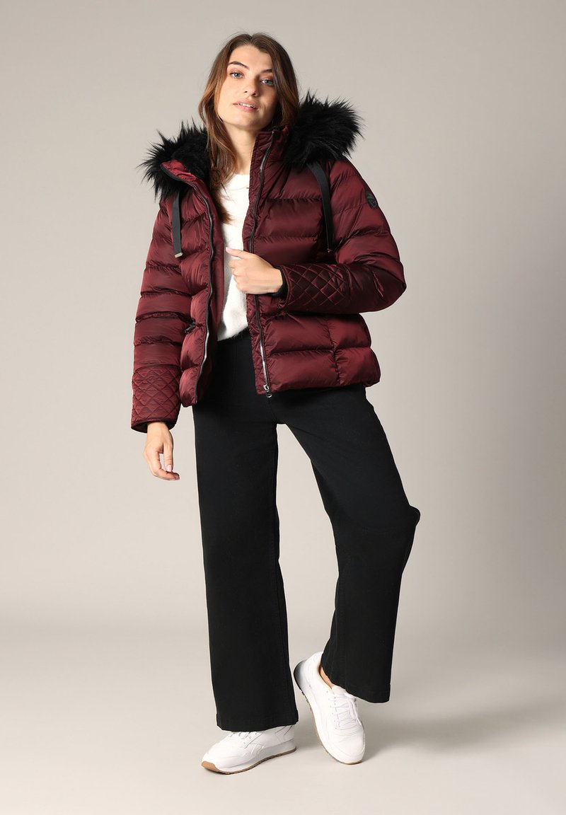 Veste matelassée bordeaux avec un motif en matelassage et une capuche en fourrure noire amovible, associée à un pantalon noir à jambes larges et des baskets blanches.