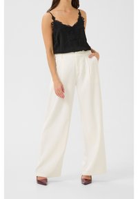 Černý saténový top na ramínka s krajkovými detaily, kombinovaný s bílými širokými kalhotami. Modelka má na sobě vínové lodičky s špičatou špičkou. Jednoduché pozadí.