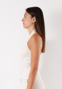 Top senza maniche, aderente, in tessuto beige chiaro con una superficie liscia. Presenta un collo alto e cuciture sottili lungo le cuciture.