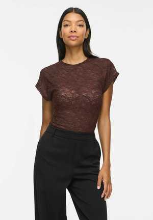 Femme portant un haut en dentelle marron à manches courtes et un pantalon noir taille haute, debout sur un fond blanc.