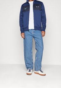 Marinblå zip-jacka med mörkblå bröstpanel, vit text "AIR," kombinerad med ljusblå denimjeans och vita sneakers med marinblå detaljer.