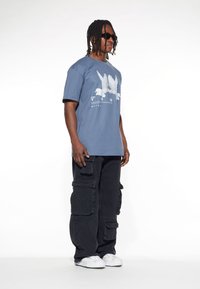 Blå bomullströja med vit fågelgrafik och text, i kombination med vida svarta cargo-byxor med flera fickor. Vita sneakers på fötterna.