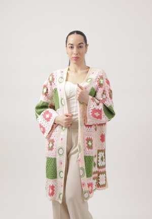 Femme aux cheveux foncés attachés en arrière portant un long cardigan crocheté coloré avec des motifs carrés roses, verts et blancs, sur un haut blanc et un pantalon beige.