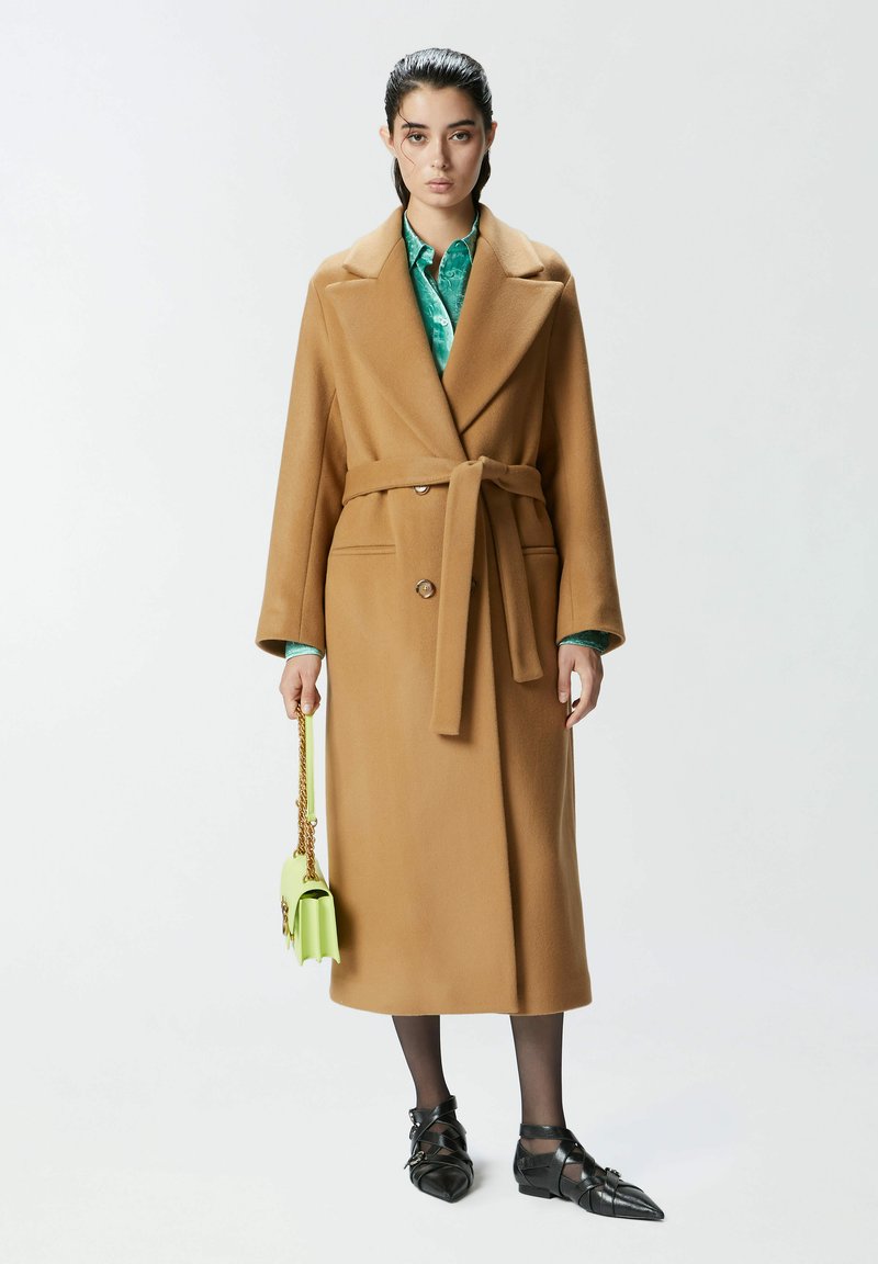 Smeđi kaput od vune s remenom u struku, dvostrukim kopčanjem i velikim reverima. Uparen s zelenom torbom i crnim cipelama.