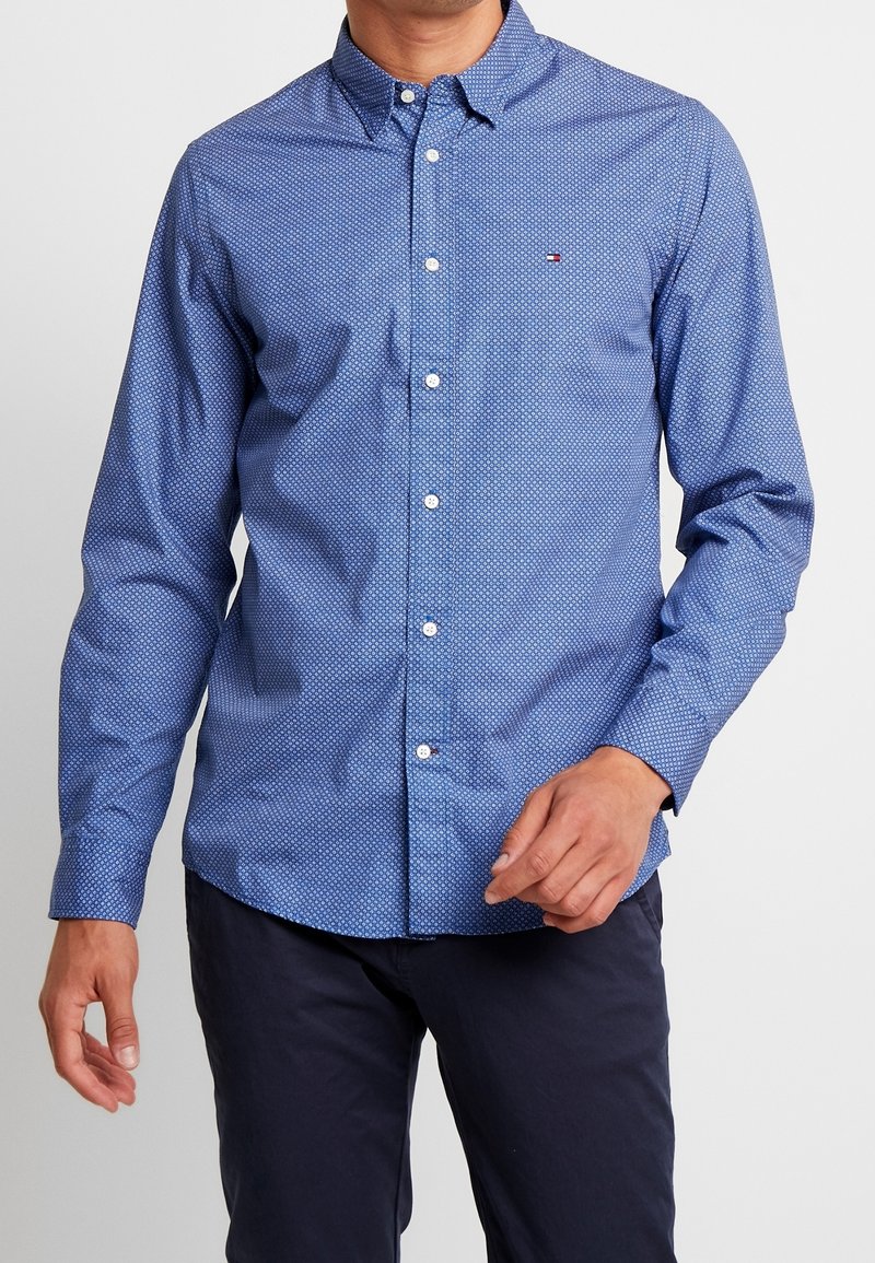 Homme portant une chemise bleue à manches longues boutonnée avec un petit motif géométrique et un pantalon foncé sur un fond uni.