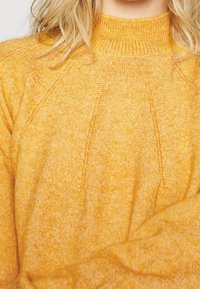 Woman wearing a soft, yellow, knit sweater with ribbed high neckline and subtle textured vertical patterns.