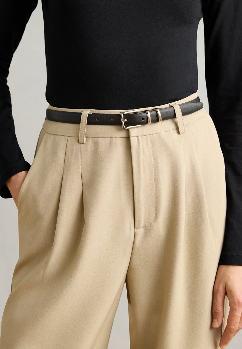 Top negro de manga larga combinado con pantalones beige de tiro alto y con pliegues, asegurados con un cinturón de cuero oscuro que presenta una hebilla plateada.