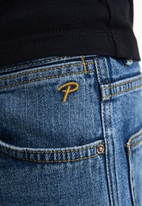 Jeans in denim blu con cuciture dorate e un piccolo logo "P" sulla tasca posteriore. Il tessuto ha una finitura testurizzata classica.