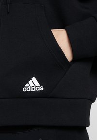 Felpa nera con cappuccio regolabile e tasca a marsupio. Presenta il logo Adidas bianco ed è realizzata in un tessuto morbido con una superficie liscia e polsini a costine.