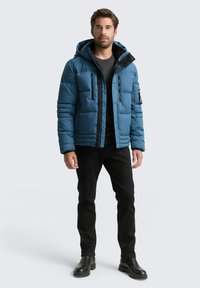 Blauer Pufferjacke mit Kapuze, schwarzen Akzenten, zwei Reißverschlusstaschen und strukturierter Oberfläche, getragen mit einem schwarzen Oberteil und einer schwarzen Hose.