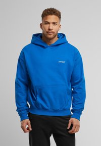 Sudadera con capucha azul, con bolsillo frontal, puños y dobladillo acanalados. Logo dorado "UPSOALE" en el pecho. Textura suave y corte casual.