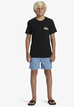 T-shirt nera a maniche corte con un logo giallo sul lato sinistro del petto, abbinata a pantaloni corti azzurri chiari e infradito nere.