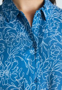 Blå skjorte med et blomstermønster i hvitt, med klassisk krage og knappeåpning. Stoffet ser glatt og lett ut.