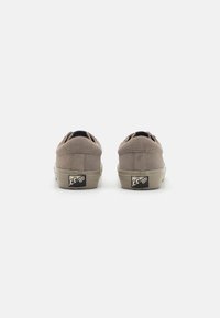 Sneaker beige in suede con retro arrotondati, suole in gomma e un'etichetta nera sul tallone. Design minimalista senza lacci visibili.