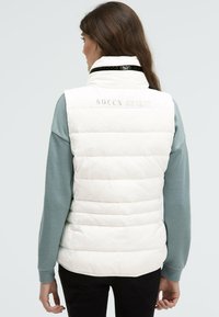 Weiß gesteppte Weste mit hohem Kragen, auf der Rückseite prangt der Aufdruck "SOCCX SPIRIT", getragen über einem helltürkisfarbenen Langarmshirt.