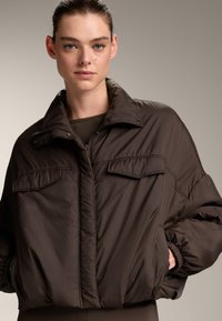 Brown puffer jacket with a high collar, two front pockets, and elastic cuffs. Smooth texture, slightly oversized fit, with a zipper closure.