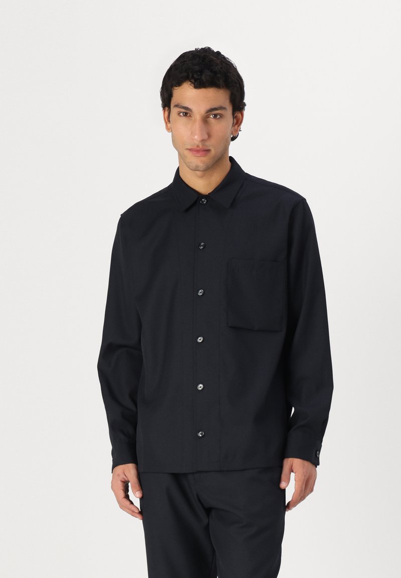 Black long-sleeve shirt with a button-down collar, front pocket, and contrasting buttons. Made of smooth fabric with a tailored fit.