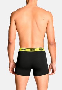 Black boxer shorts with a yellow Puma logo waistband. The fabric appears smooth, fitting close to the body, with a minimalist design.