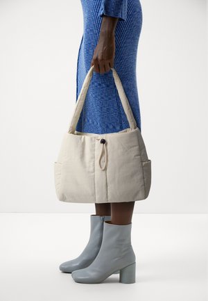 Personne en robe bleue tenant un grand sac matelassé beige près de bottines gris clair à talons carrés sur fond blanc.
