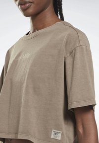 Hnědá tričko s krátkým rukávem, vyrobené z měkké bavlny, má kulatý výstřih a etiketu Reebok na lemu; přírodní barvivo s hladkou texturou.