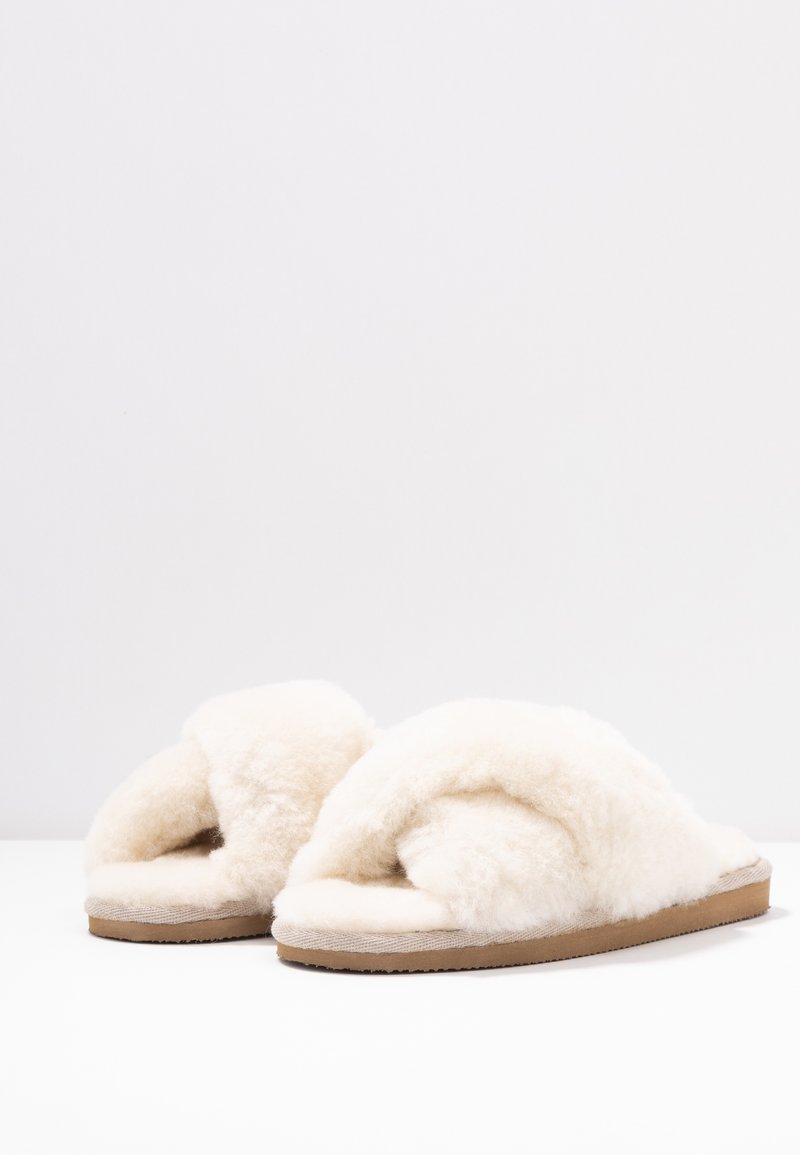 Een paar crèmekleurige, fuzzy open teen slippers met een bruine zool op een witte ondergrond tegen een lichtgrijze achtergrond.