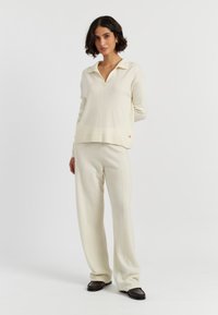 Cremfarbener Strickpullover mit Kragen, kombiniert mit passenden weit geschnittenen Hosen. Das Outfit hat ein dezentes rotes Logodetail auf dem Pullover.
