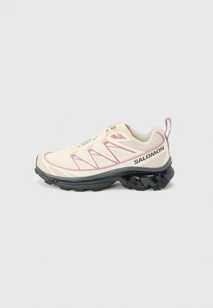 Off-white Salomon trailrunningschoen met roze accenten, zwarte robuuste zool en veterontwerp tegen een effen lichte achtergrond.