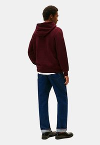 Felpa bordeaux con cappuccio regolabile, vestibilità rilassata, abbinata a jeans blu dritti e scarpe nere. Dettagli di cucitura visibili.