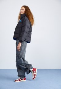 Czarna kurtka puchowa z wysokim kołnierzem, niebieską podszewką, szare spodnie cargo i czerwone sneakersy Nike z białymi akcentami.