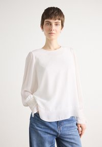 Witte blouse met lange mouwen en een ronde halslijn, voorzien van transparante mouwen. Textuurmanchetten hebben een beadaccent. Gepaard met blauwe jeans.